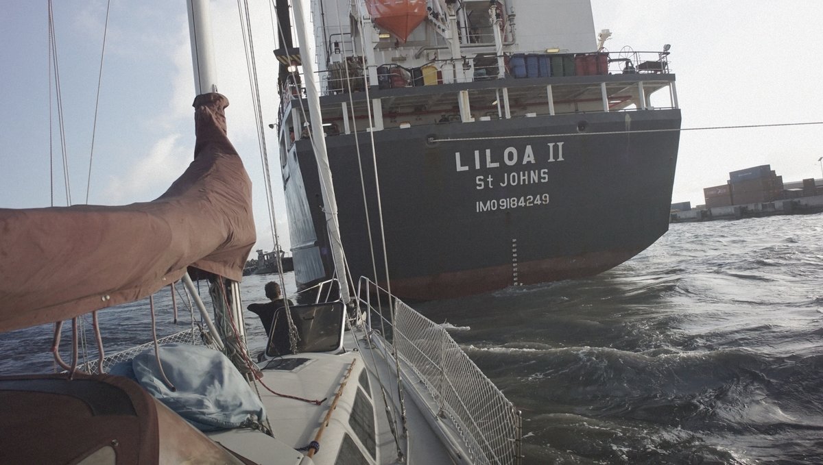 Rek sitting on the bow of a sailboat while moored into a port with the stern of a large cargo ship just a few short feet away as it is trying to dock, its powerful propellers raising wakes