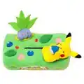 A tissue box cover designed to look like grass, with a plush Oddish sitting and a plush Pikachu lying on top; Pikachu is holding a Chesto Berry; other berries have been added as patches onto the plushy fabric grass.