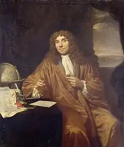 A man wearing a long, curly wig and a full robe is sitting, looking out. His left arm rests on a small table, with his left hand holding a box. Behind him is a globe.