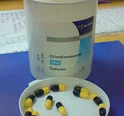White bottle on blue pad atop a desk. The bottle cap is off, and is upside down on the pad in front of the bottle. In the cap are a dozen black-and-yellow capsules.