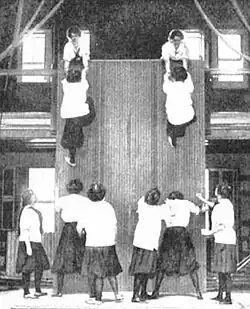 Students co-operate to scale a high wall. The activity involves lifting someone up, wall walking, and pulling someone up; c. 1900.