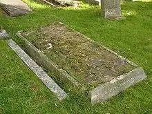 A horizontal gravestone, badly dilapidated and with grass growing among the cracks