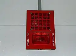 a red device built into a wall.  The top half is a speaker and the bottom half is a red light