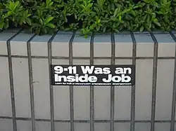 A photo of a black, rectangular sticker that falsely claims that "9/11 was an inside job" in big white text, under the big text was a suggestion to go to three conspiracy theory sites.