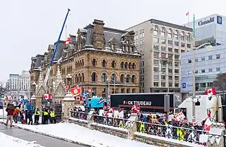 Crowds of convoy protesters gathered outside the Office of the Prime Minister and Privy Council Building (which is located across from Parliament Hill). Many protesters are waving Canadian flags or holding signs with slogans like "wake up" and "my body, my choice". A truck is parked next to the office building so as to block the road to Parliament Hill. The truck has the text "Fuck Trudeau" painted on its side, except that the U in "Fuck" is replaced with a maple leaf exactly like the one on the Canadian flag.