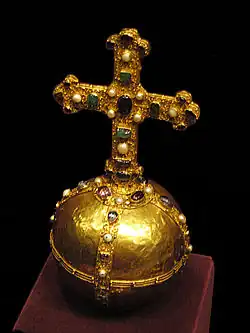 A smooth gold sphere wrapped in raised bands along the equator and two perpendicular longitudinal rings. A cross, taller than the orb's diameter is mounted on top. The bands and cross are garishly embossed with a curly vine pattern and studded with gems.