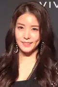 Woman with long, wavy dark brown hair, smiling at the camera. She is wearing large hoop earrings and is dressed in a black outfit.