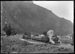 A steam locomotive hauls a train of goods waggons. It is passing a settlement of wooden buildings. In the near background is a steep mountain side.