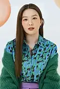 Woman with long, straight brown hair, wearing a colorful, patterned blouse in shades of blue and green, paired with a fuzzy green cardigan. She has a calm, composed expression and is facing the camera.