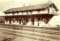 A black and white photograph of a two-story train station, with multiple railroad tracks in the foreground