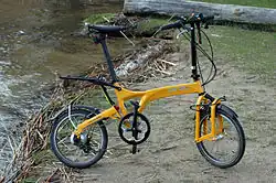 A yellow 2011 Birdy full suspension folding bicycle with a leading arm anti-dive front suspension and a hinged rear swingarm suspended by an elastomer standing in green nature near a river.