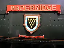 A 'West Country' class enamelled metal nameplate and shield mounted on flat metal casing covering the locomotive's boiler. The nameplate comprises a scroll, and below this is a shield containing a picture of a coat-of-arms. A second scroll is below the shield, allowing identification as a member of the 'West Country class'.