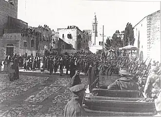 Towers and buildings some with people looking down from roofs with local people and soldiers in a large square with motor car in foreground