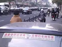 A car parked in a bike lane adjacent to a busy street in Shanghai. The lane is separated from the street by a fence. A sticker on the car reads "Embrace the world expo while observing the law, Driving with courtesy for a good city image."