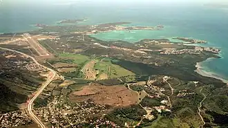 an aerial view of the island's coast, with buildings and streets in the bottom left, open greenspace in the middle, a highway on the left. A large body of blue-green water surrounds the top of the island, as well as on the right side.