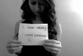 A black-and-white image of a girl in her mid-teens holding a cut-out piece of paper or card with the words "I have nobody, I need someone" and a sad emoticon written on it.