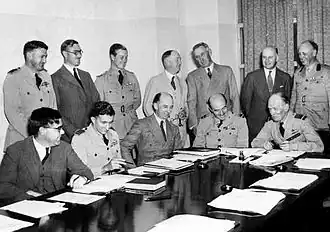 A dozen men behind a large table, half of whom are in military uniform and half in civilian clothes, seven standing and five seated