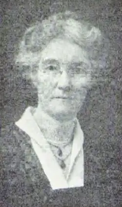 An older white woman with grey hair in a bouffant updo, wearing glasses and two pendants.