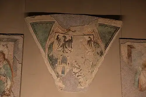 Triangle on which a shield is painted. The latter is divided into four. On the first part on the upper left there is a black bird on a white background, the same on the upper right but erased, on the lower left there is a white castle on a blue background and the last drawing is barely visible.