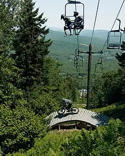 A mountain biker rides down a wooden ramp feature while two other bikers ride a chairlift overhead at Mt. Abram. The background shows green forest and distant mountains.