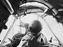 Pilot in a small cockpit. He is wearing a parachute and leather helmet. This is a still from movie footage shot by Damien Parer.