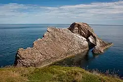 A large brown rock with a striking shape lies just beyond a rock coast in a blue sea under a pale blue sky. The left hand and foreground part of the rock is wedge shaped and the sedimentary rocks it is made off are set at a 45 degree angle to the horizontal. A second part of the rock is in the shape of an arch with a thick top section and a thin downward leg. The whole structure has a strangely contrived air, suggestive of a wrecked ship.