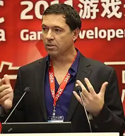 Portrait of a White man wearing a black jacket in front of a podium with two small goose-neck microphones. His hands are up near his chest while he speaks and looks to the left.