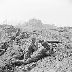 Five men with two guns in a devastated landscape. The remains of a bombed town are in the background