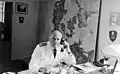 Black-and-white photo of a man wearing a white military uniform sitting at a desk, making a phone call.
