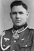 The head of a young man, shown in semi-profile. He wears a military uniform with a military decoration in shape of an iron cross displayed at the front of his shirt collar. His hair is parted and combed to back.