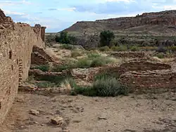 A partly overcast sky and subdued sunlight over a roughly six-foot tall wall of dusky tan sandstone bricks that vary somewhat in size. The wall runs diagonally from the immediate foreground at left toward the right, running perhaps several dozen feet to the near middle distance. A few feet to the right, in the middle foreground, a low ring of similar blocks delimits a circular pit sunk into the ground. The remains of several other ruinous low walls, perhaps one to three high at most, are arrayed in parallel; they align left to right from the high diagonal wall. Perhaps a mile distant to the center and right, a canyon wall slopes gradually level to meet the valley floor on which the walls sit.