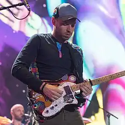 Buckland on stage with his guitar, wearing a black hat and a shirt with colourful details