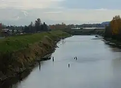 A stream perhaps 50 feet (15&nbsp;m) wide flows between raised, grass-covered banks on either side. Several poles or sticks, perhaps remnants of a derelict structure, poke above the water in places. Low buildings and many trees are in the middle distance. A mountain range dominated by a snow-covered peak is visible in the far distance.