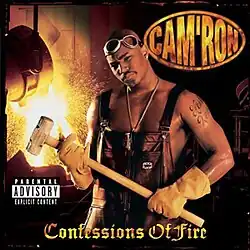 An image of Cam'ron wearing overalls but no shirt. He has googles on his head and is wearing heavy worker gloves to hold a sledgehammer. He appears to be in a smeltery or foundry and in the background a vat is pouring molten metal, causing sparks to fly.