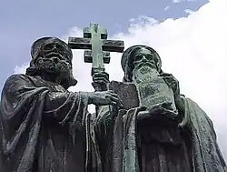 A photograph of statue, which depicts two bearded and hooded men; the one on the left is holding up a cross and the one on the right is holding up a book