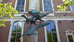 Two Deinonychus figures balance on a claw-like form, in active poses as if they are pursuing prey. In the background is a banner advertising The Academy of Natural Sciences, and the brick structure and windows of the museum itself. It's late in the afternoon on a sunny day in July, and a sunbeam striking the face of one of the sculpted dinosaurs casts dramatic shadows.