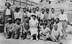 A crowd of mostly bearded Greek workmen, with a woman in a white dress and sun-hat seated in the centre.