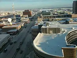 Snow-covered flat roofs and snowy streets seen from slightly above. In the far background is a frozen lake and some low hills