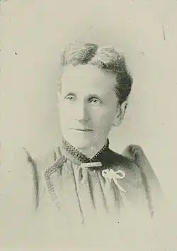 B&W portrait photograph of a middle-aged woman with hair in an up-do wearing a high-collared blouse with a white ribbon pinned on the left side.
