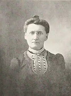 B&W portrait photo of a middle-aged woman with her hair in an up-do, wearing a dark blouse with a frilly front.