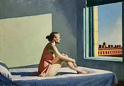 A woman sits on a bed gazing out of a sunlit window