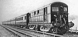 A black and white image of an electric locomotive hauling at least 6 coaches, shown with the electric locomotive on the right. A track in the foreground is electrified with the fourth rail system. The locomotive is shown with two pick-up shoes.