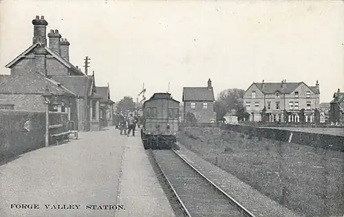 A black and white image of a train static in a station, with the platform on the left and houses beyond on the right