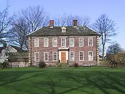 A two-storey brick-built mansion in its own well-kept garden of green grass. The ground floor has six windows – three on each side of a wooden front door. The second floor has seven windows. The house has a grey slate roof surmounted by two small brick chimneys. Behind the house are leafless trees and behind those, a clear blue sky.
