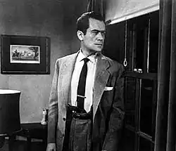 Black and white film still of Frank Lovejoy in character, wearing a suit and tie, looking surreptitiously out of a window