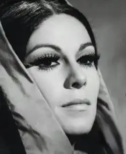 Three-quarter profile black and white glamour publicity shot of an unsmiling young woman in heavy makeup and false eyelashes, and long, straight, dark hair tied up in a bun, with centre parting. She is wearing a hood.