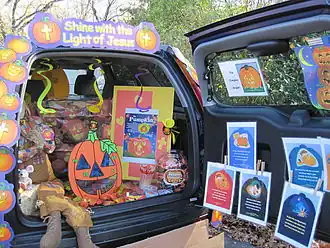 An open trunk of a van decorated with paper jack-o'-lanterns. Inside the trunk is a large jar full of candy and above the vehicle is a paper sign with the words "Shine with the Light of Jesus".