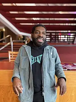 A photo Hanif Abdurraqib wearing a denim jacket over a black hoodie looking off camera and leaning against a counter.