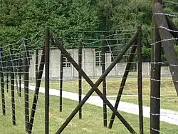 Taken from outside the reconstruction of a barracks, the photo shows a barbed wire fence, and beyond it a grassy area with a small timber hut