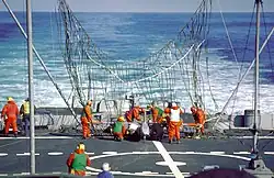 A large net on the back end of a ship. Several orange clad crewmen are working to free a white singed object from the net.